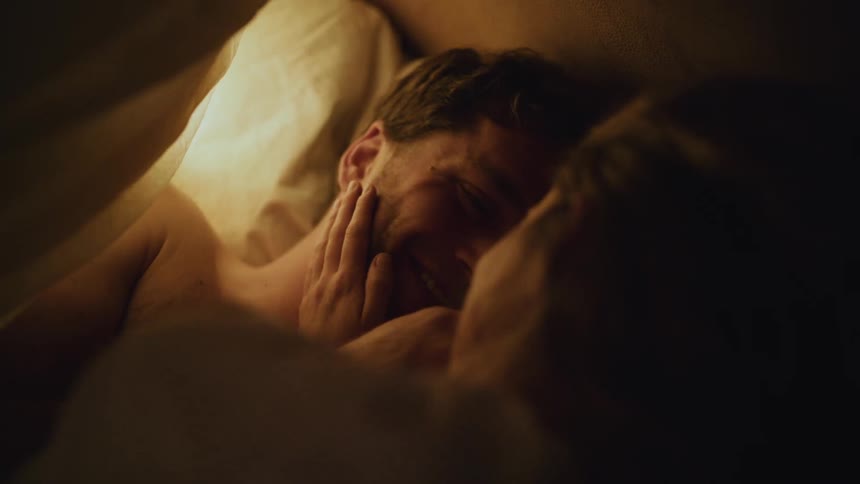Happy romantic young couple lying in the bed covered with a blanket caressing and talking to each other