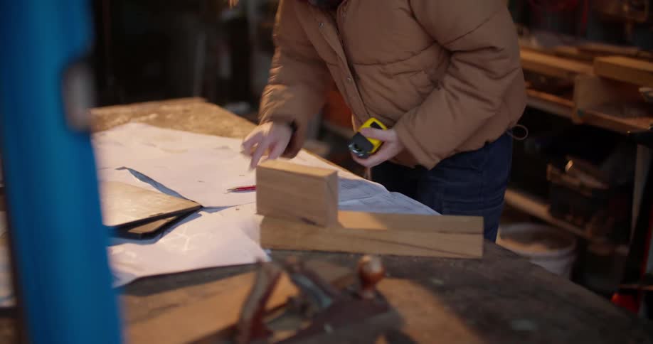 Woman in workshop measures wooden boards and blocks and writes numbers down on paper