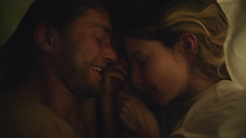 Couple lies together on bed holding hands and looking into each others eyes