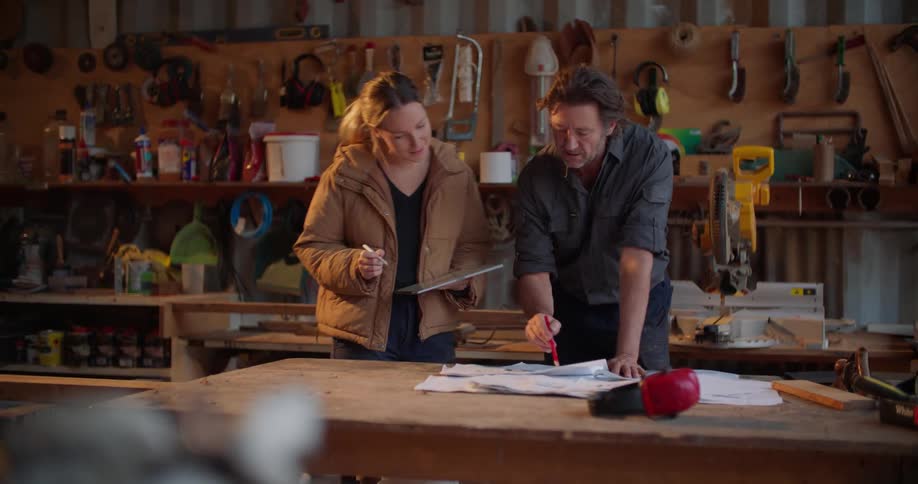 A woman carrying a tablet walks up to a man looking at some papers in a tool shop