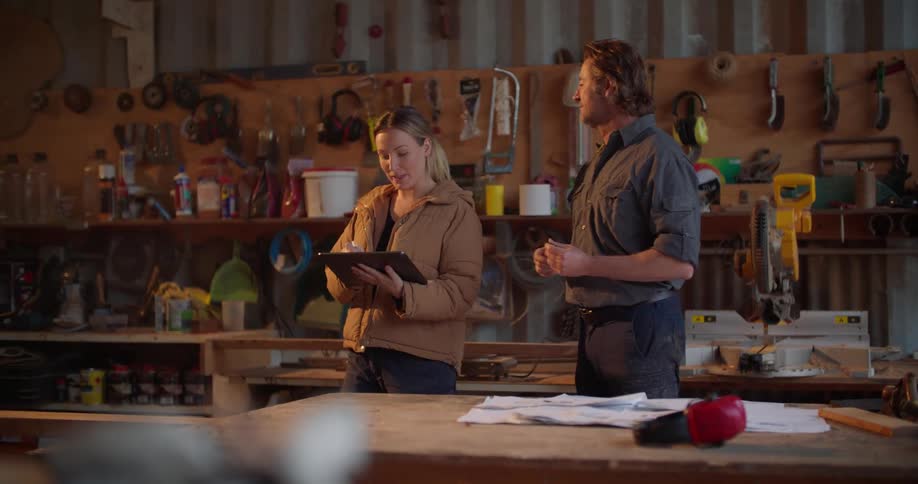 A woman holds a laptop while going over blueprints in a garage workshop