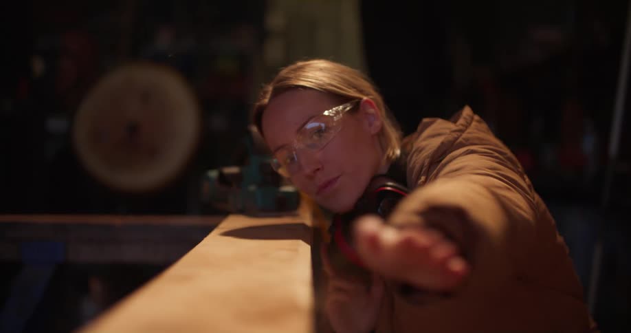 A woman squats down to see how smooth some wood is then blows the sawdust off and stands up