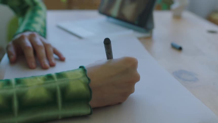 An artist sitting at a table drawing on paper inspired by a womans face on her laptop