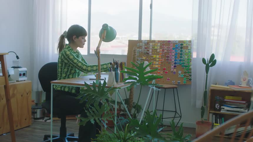 A woman walks to her desk carrying a laptop and sits down with a color swatch board and paintbrushes on display