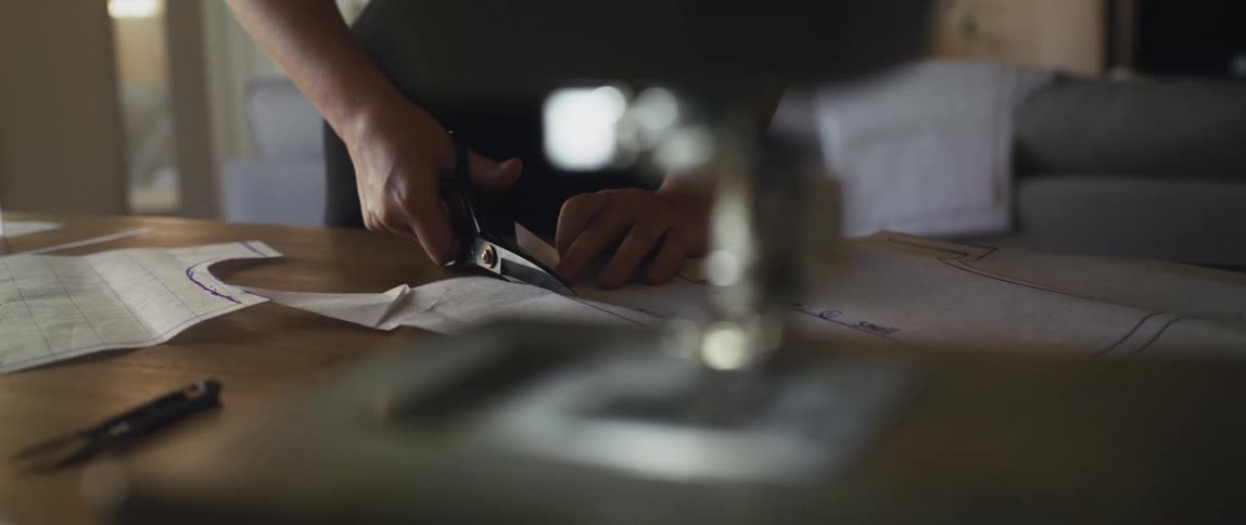 A man standing at a worktable near his sewing machine cutting out a pattern