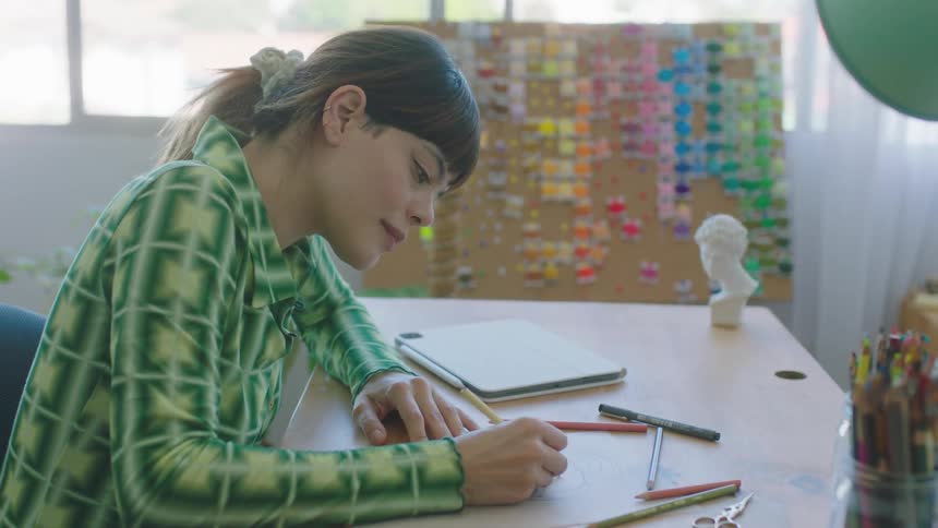 A woman sits at a desk and draws after looking at a colorful board for inspiration