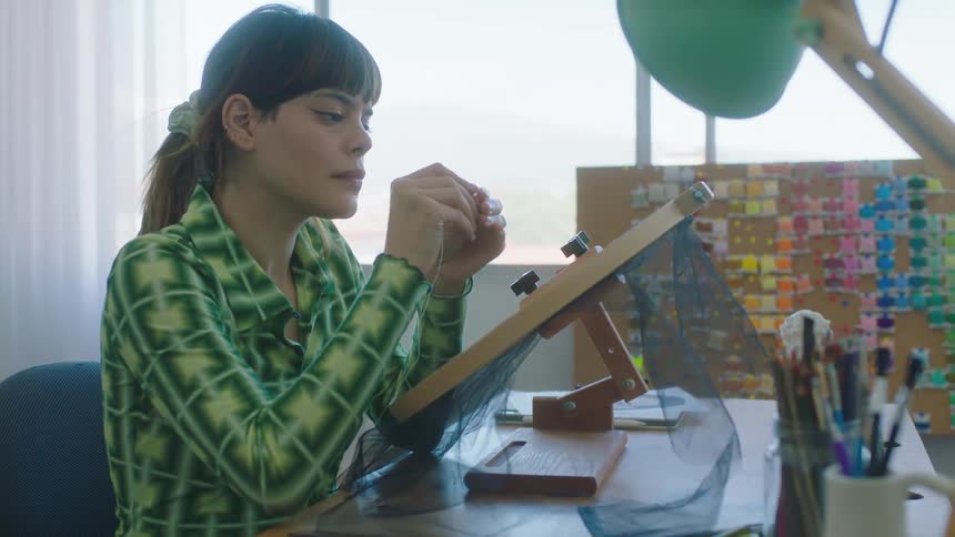 A woman threads something around her finger while working on a project in front of her