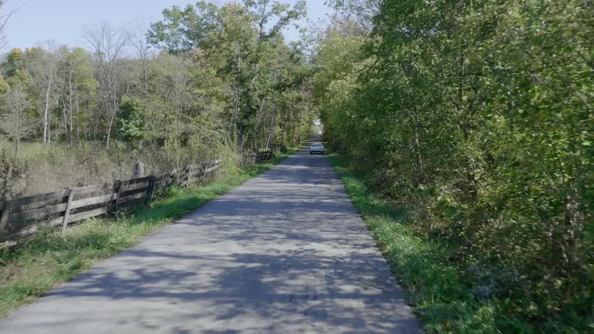An older white car travels down a country road lined with trees and a wood fence
