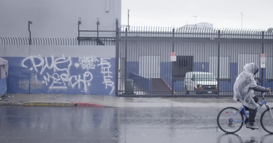 A man wearing a plastic rain poncho rides a bike down the street in the rain