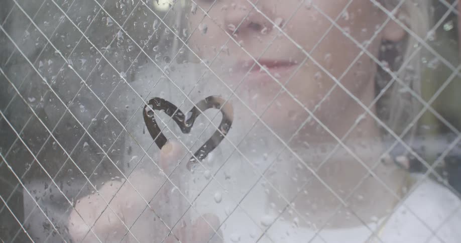 A woman blows on a window and draws a heart in the fog her breath created
