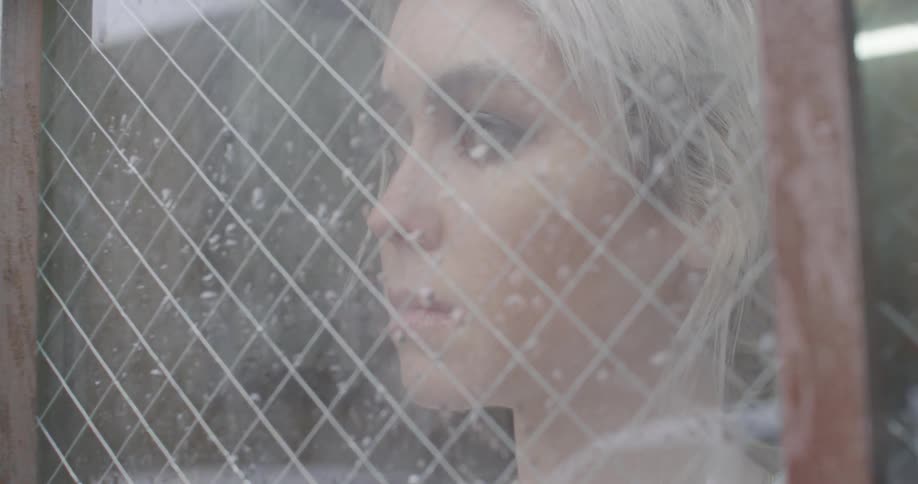 A woman looks through a security glass window that has droplets of water on it