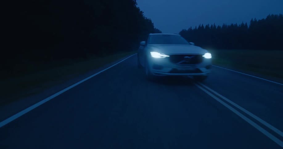 A white automobile with headlights on is traveling fast on a road and moves to the next lane