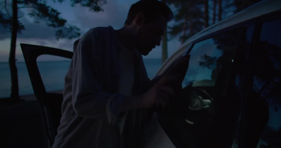 Man steps up to parked car sitting with the drivers door open and taps on the back door with the water in the background