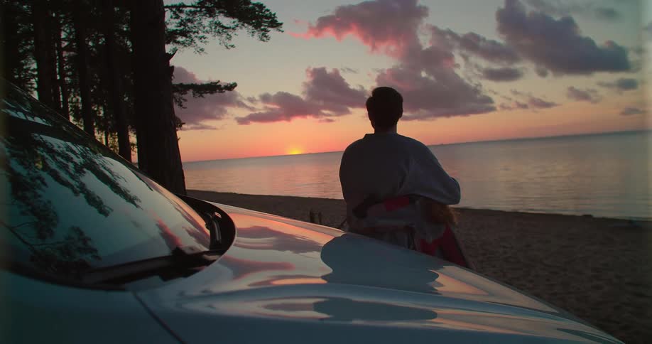 A woman runs into the arms of a man leaning against a car on the beach
