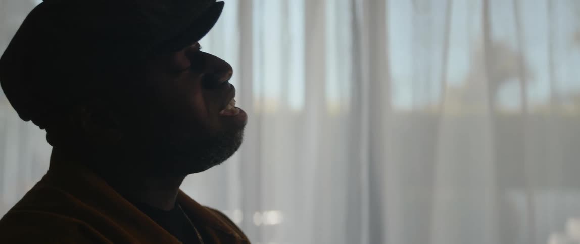 Male musician with black cap and orange jacket plays piano singing song in room