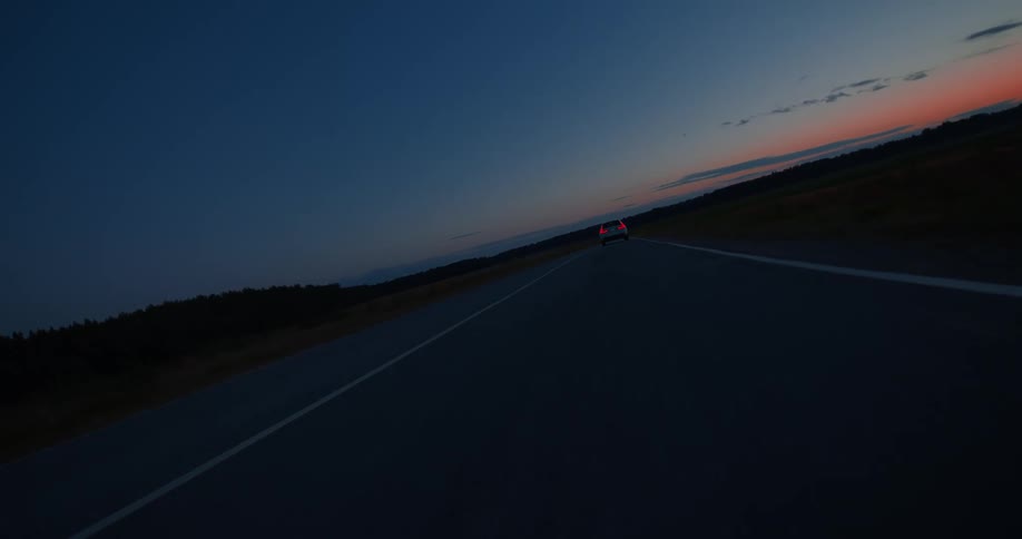 The tail lights of a white automobile are lit as it travels down the road at dusk