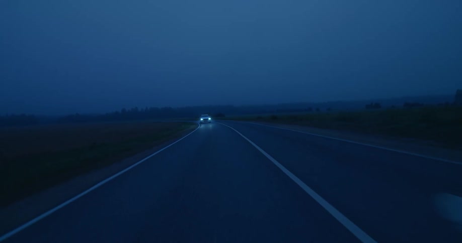 An automobile with the headlights on is traveling on a road at dusk