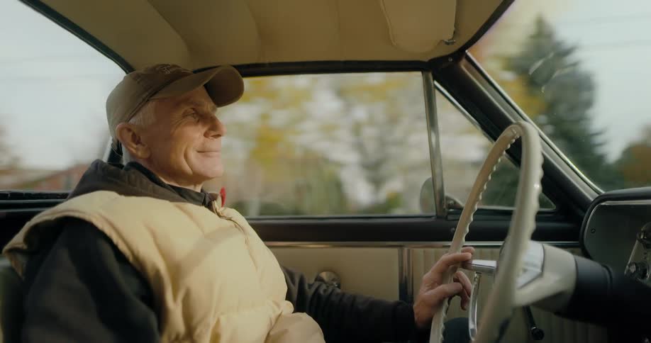 An elderly man wearing a cap and a coat is driving an automobile through a neighborhood