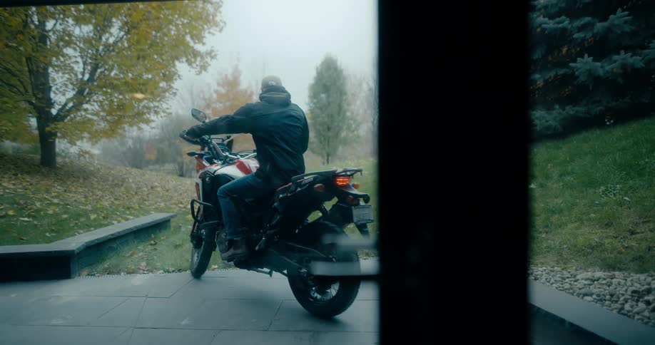 A man wearing a coat gets on a motorcycle and takes off through a field with trees in fall colors