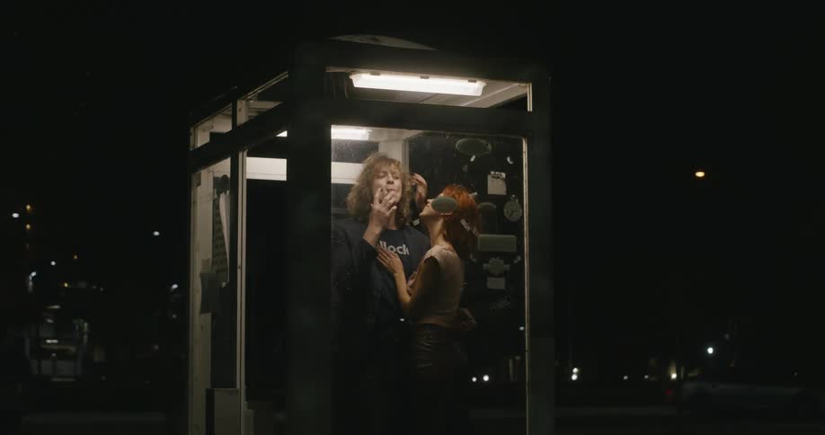 A woman plays with her boyfriends hair while they share a cigarette in a lit booth at night
