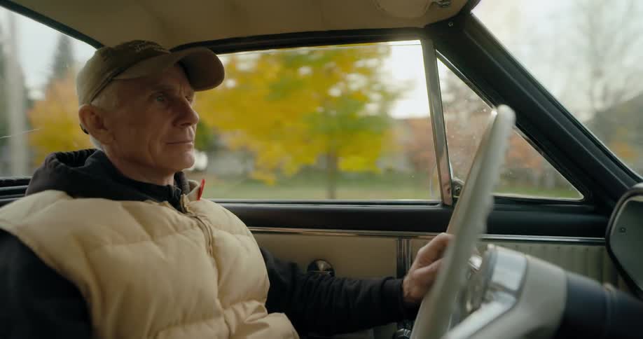 An elderly man wearing a cap and a coat is driving an automobile through a neighborhood