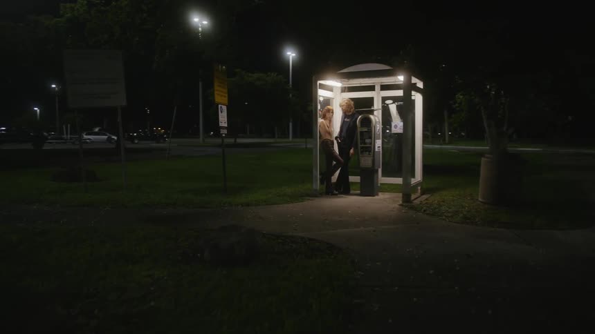 A man and woman standing in a parking lot kiosk at night smoking and flirting