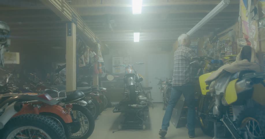 White haired man picks up and places object on shelf before picking up tool near motorcycle
