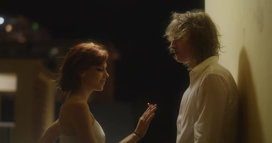 A man leaning against a wall outside sharing a cigarette with his girlfriend caressing and kissing her