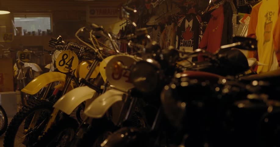 Row of racing motorcycles sit next to clothes with graphics on them in storage area