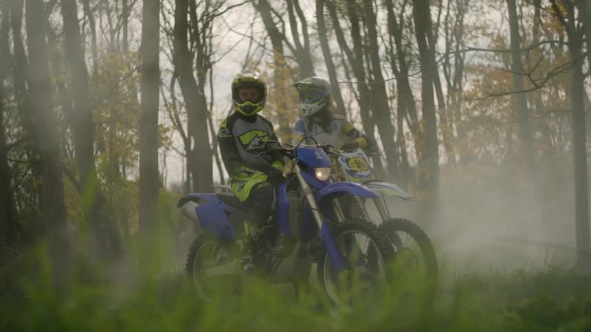 Two teenagers wearing helmets are sitting on motorbikes in a foggy forest