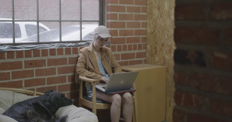 A woman sits in a chair working on a mac laptop while a dog sits near her
