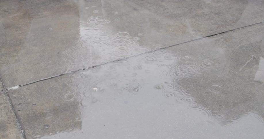 Raindrops fall into puddled water on a sidewalk