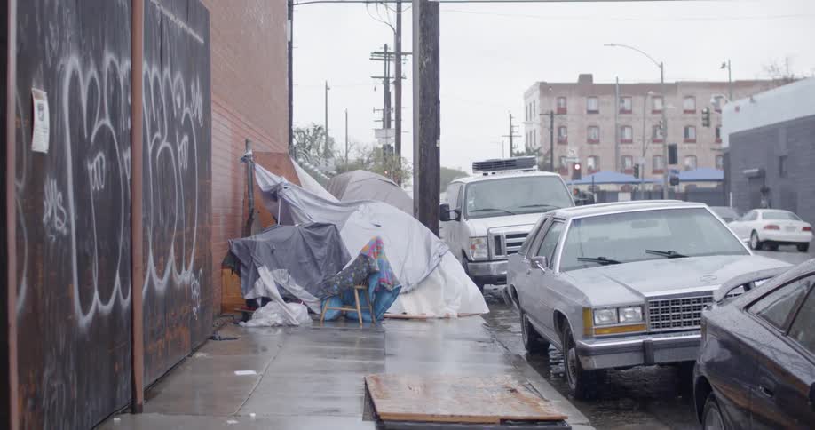 Rain falls on a homeless encampment on a sidewalk in an urban area