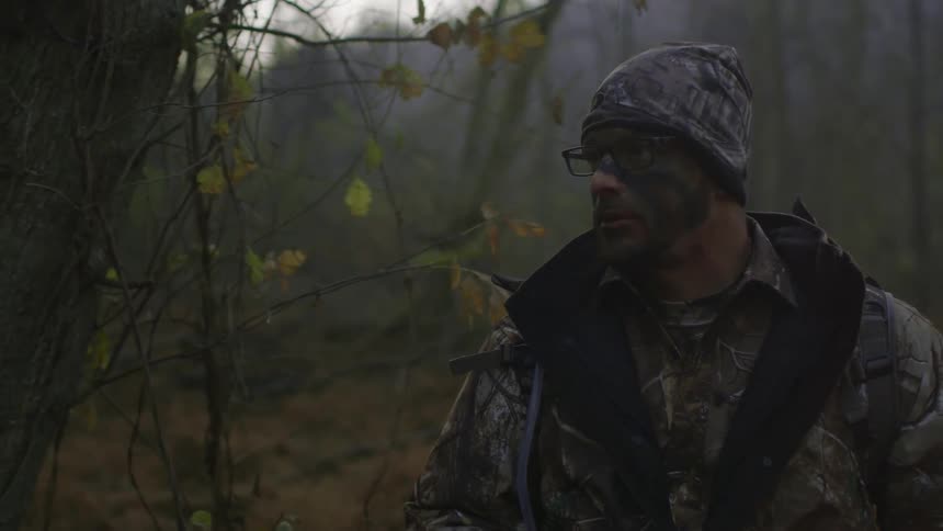 A man wearing camo and carrying a bow is walking through the forest