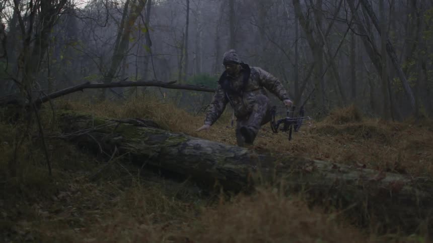 A man wearing camo and carrying a crossbow climbs over a fallen tree