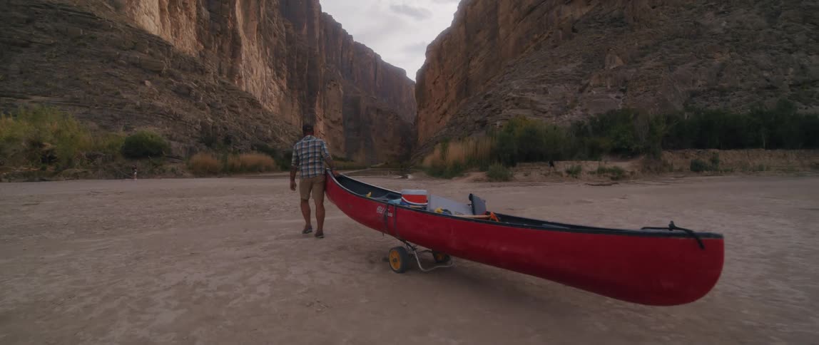 A man pulls a canoe mounted on a dolly through a field towards the entrance of a large canyon