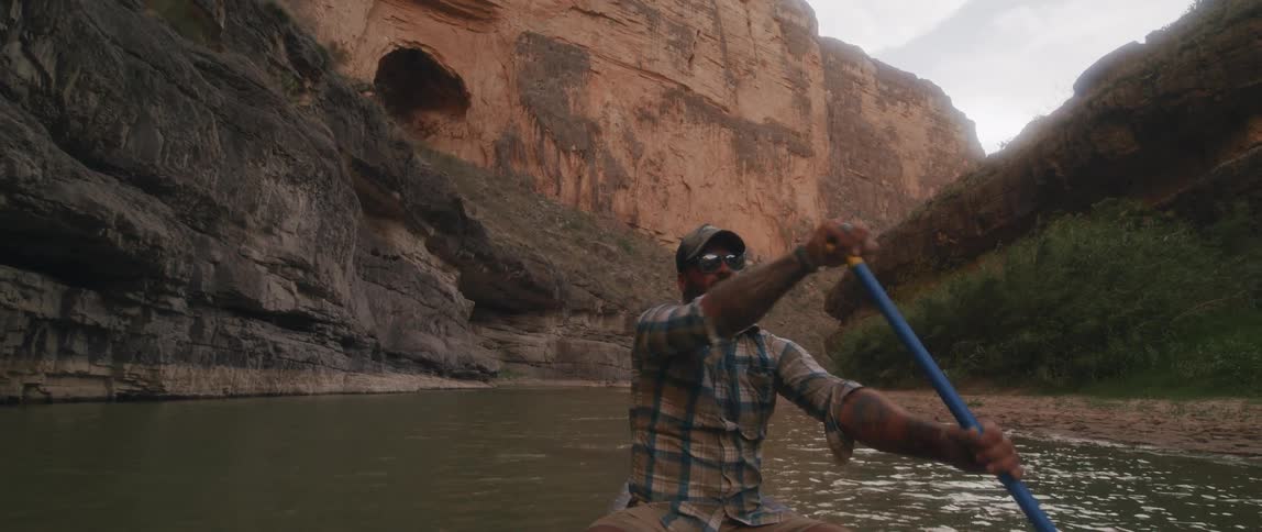 A man slowly paddles a kayak on a river in a canyon on an overcast day