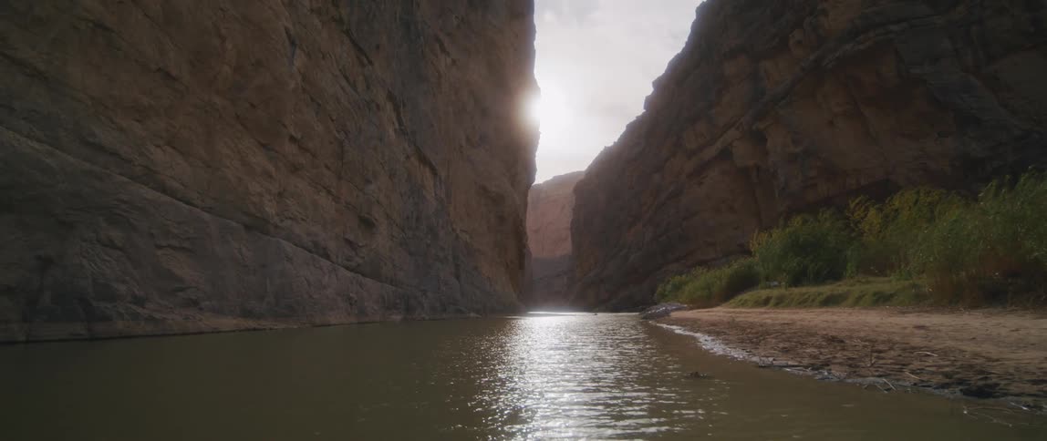 A small piece of land with plants along a canyon river as the sun shines on the ripples in the water