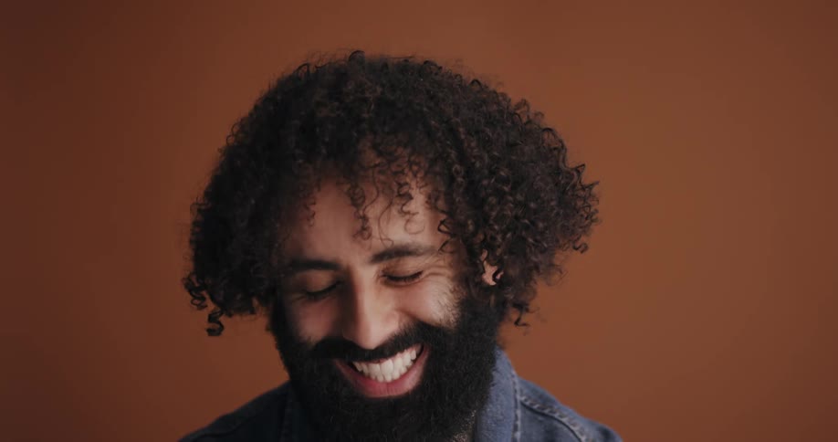 Man with curly hair and beard laughs at camera in front of brown wall