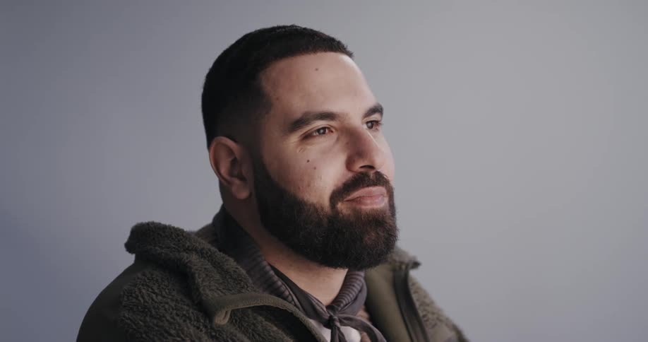 Smiling bearded white man wearing warm clothes sitting down in front of a white wall at a studio