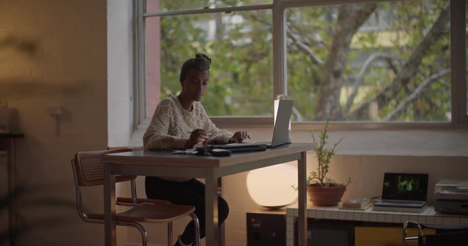 A woman uses a laptop and tablet while sitting at a table indoors during the day