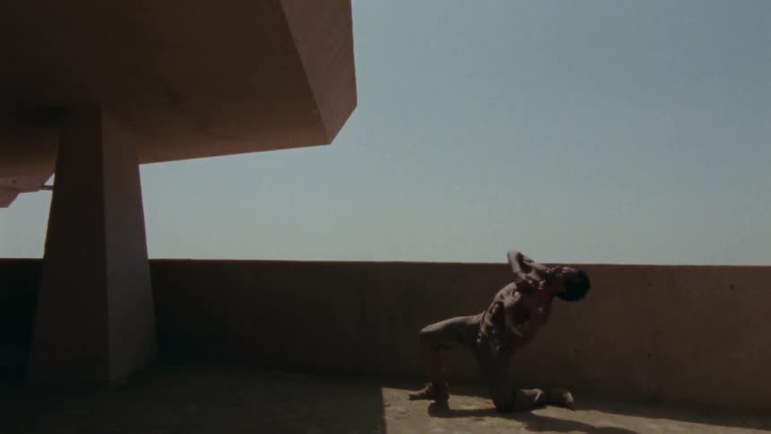 A shirtless man dances under a clear daytime sky on a large balcony