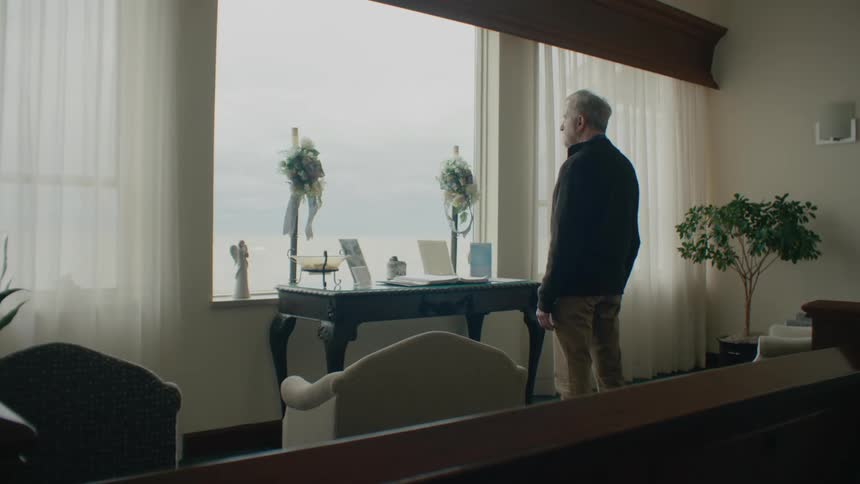 Elderly man standing in the living room looks out a window