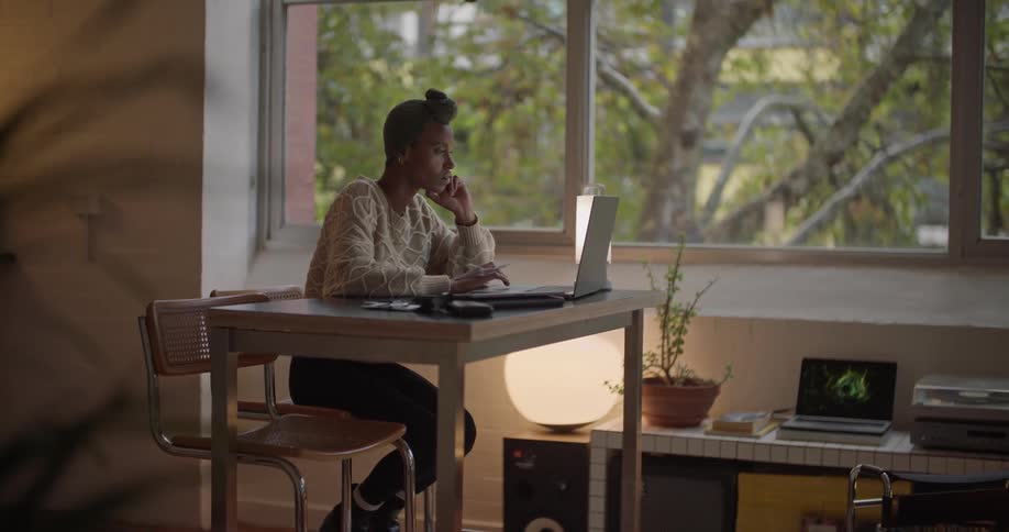Black woman wearing a head wrap sitting at the desk near the window using her laptop