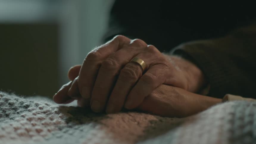 An elderly man lifts his wife's hand from the blanket and holds it in both of his