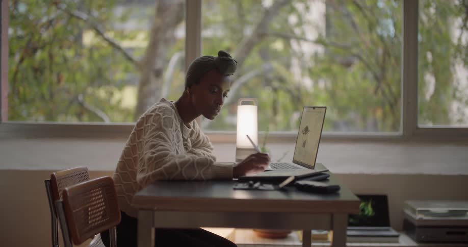 A woman is sitting at a desk working on a laptop computer and writing on a tablet