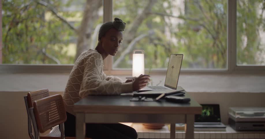 A woman is sitting at a desk working on a laptop computer and writing on a tablet