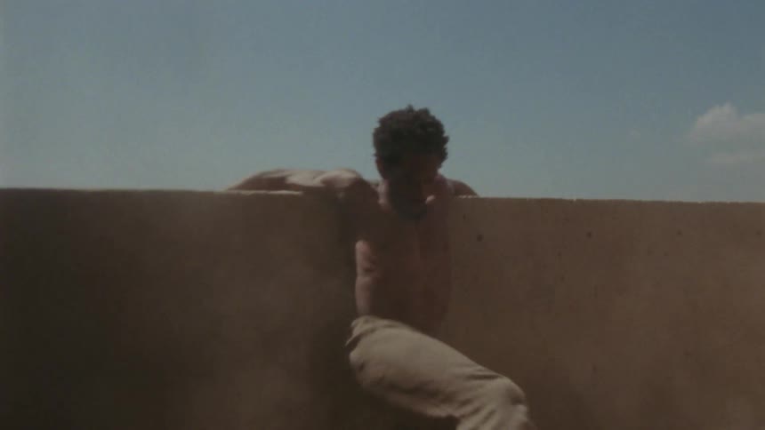 Shirtless man reclined on a wall shakes his body and legs kicking up dust and sand