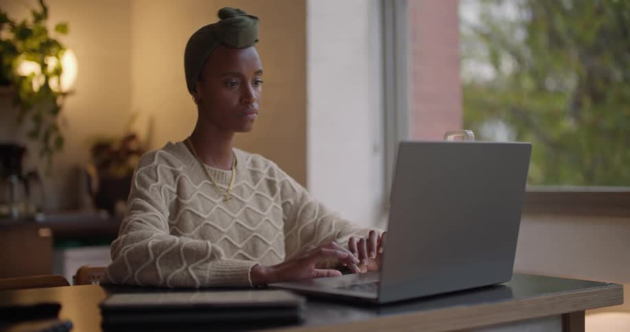 A woman is sitting at a desk working on a laptop computer and writing on a tablet