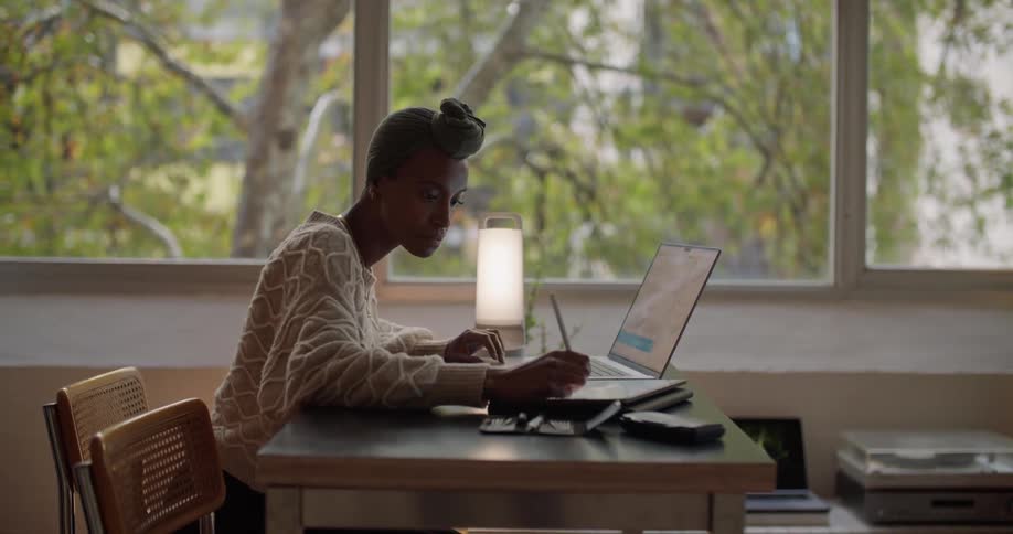 A woman is sitting at a desk working on a laptop computer and writing on a tablet